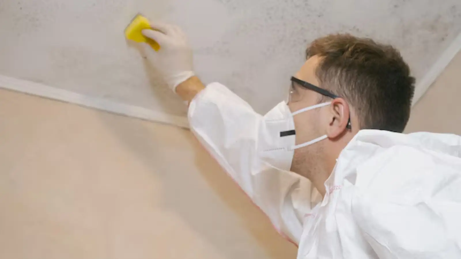 Person in a mask and protective suit wiping mold from a wall surface with a sponge